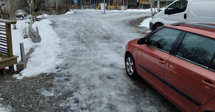 Isig uppfart med röd bil till höger och vit skåpbil i bakgrunden, omgiven av snö och träd i en byggarbetsplatsmiljö.