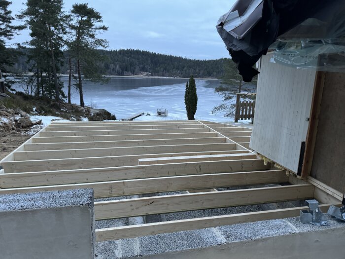 Bjälklag under konstruktion vid en sjö, omgivna av träd. Förberett för ytterväggar, med en inspektionsstege i bakgrunden.