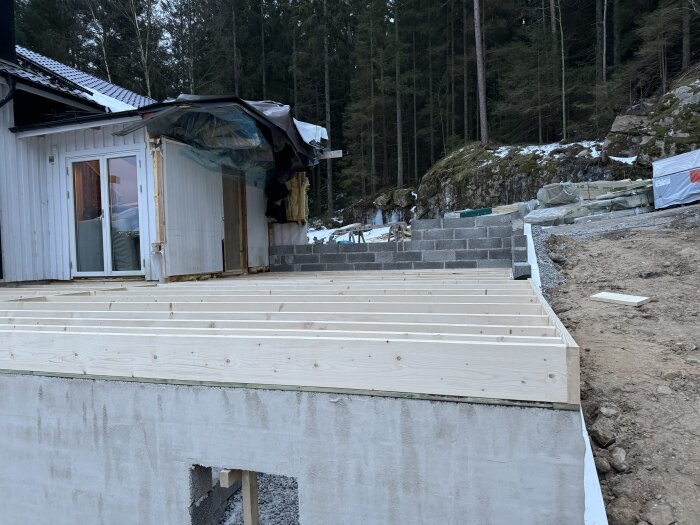 Byggplats med nytt bjälklag, halvfärdig vägg och täckt del av hus. Omgiven av skog, synlig jord och byggmaterial staplat intill.