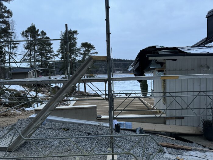 Byggarbetsplats med ställning och bjälklag framför hus nära sjö, omgiven av träd, med vinterlandskap i bakgrunden.