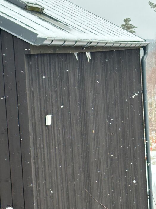 Istappar hänger från en takränna på ett plåttak, med snö och ett takfönster synligt ovanför rännans position.