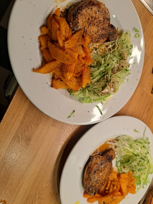 Två vita tallrikar med grillat kött, morotsstavar och riven sallad på ett träbord.