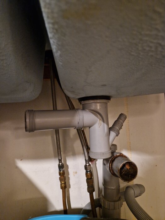 Gammalt avloppssystem under diskbänk med grå plastkopplingar och metalrör från 1977, kopplingsproblem vid köksrenovering.