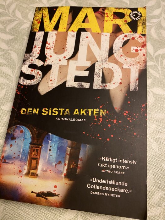 Omslag av kriminalromanen "Den sista akten" av Mari Jungstedt med blodstänk och bilder på en kvinna och en byggnadsinteriör.