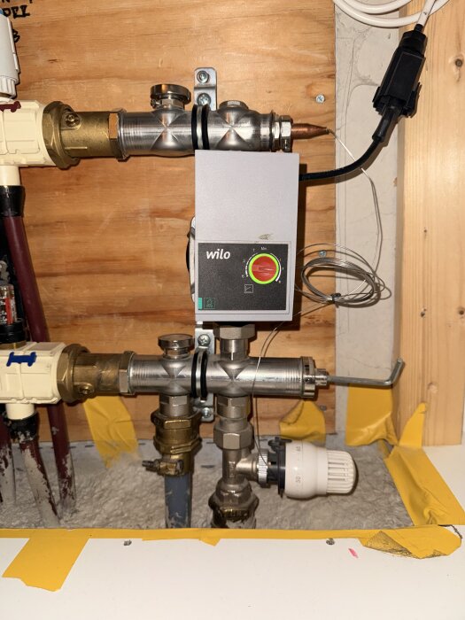 Cirkulationspump och rörkopplingar i golvvärmesystem med termostatventil och gul tejp, monterade på en träpanel i husets värmesystem.