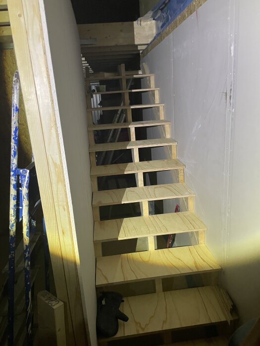 Halvbyggd trappa med elva steg och lösa plansteg. Trappen saknar viloplan och svänger till vänster efter uppgång. Byggmaterial synligt runt om.