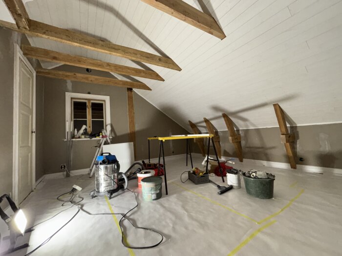 Renovering av sovrum med synliga träbjälkar, arbetsverktyg och färgburkar på plasttäckta golvbrädor.