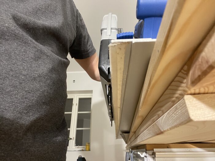 Person använder cirkelsåg på träplankor i ett rum med vitt tak och fönster i bakgrunden.