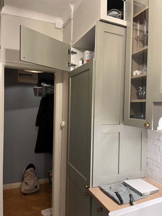 Öppen köksskåpsdörr ovanför integrerad kyl; diskproblemet diskuteras, möjligen för ventilation. Köket har grågrön skåpinredning och träbänkskiva.
