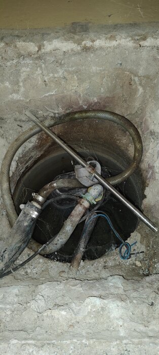 Dräneringspump i hål under betonglucka, omgiven av slangar och verktyg. Vatten synligt i botten av hålet.
