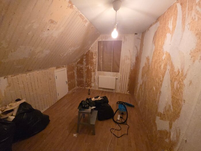 Rum under renovering med avskalade tapeter, trägolv, lampa i taket, och verktyg på en pall. OSB-skiva på en vägg och fönster täckt av papper.