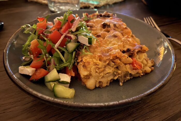 Makaronipudding med chorizofärs, tomat, champinjoner och ost, serverad med sallad innehållande fetaost, gurka och tomat på grått fat.