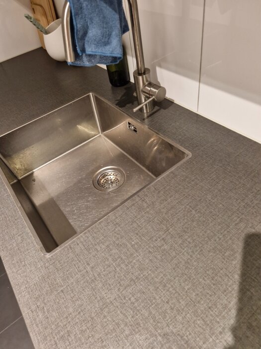 Köksbänk med diskho i rostfritt stål, grå bänkskiva, kran med blå handduk, fläckar synliga nära diskhon.
