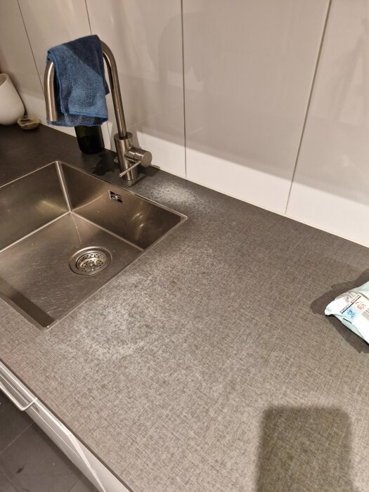 Grå bänkskiva i kök med fläckar runt diskhon, blå handduk på blandaren, vita kakelplattor i bakgrunden.