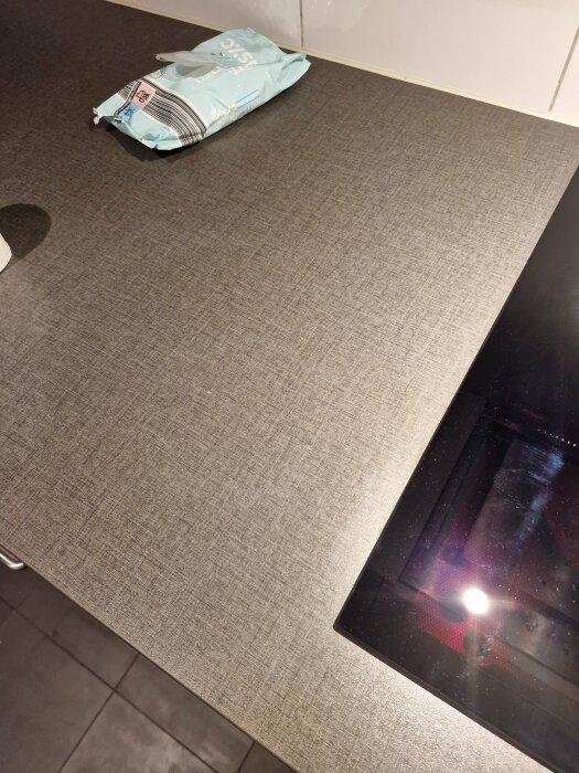Grå Ikea bänkskiva med tydlig linjemönster, en del av en spis syns till höger. En vit tub och en påse på bänken.