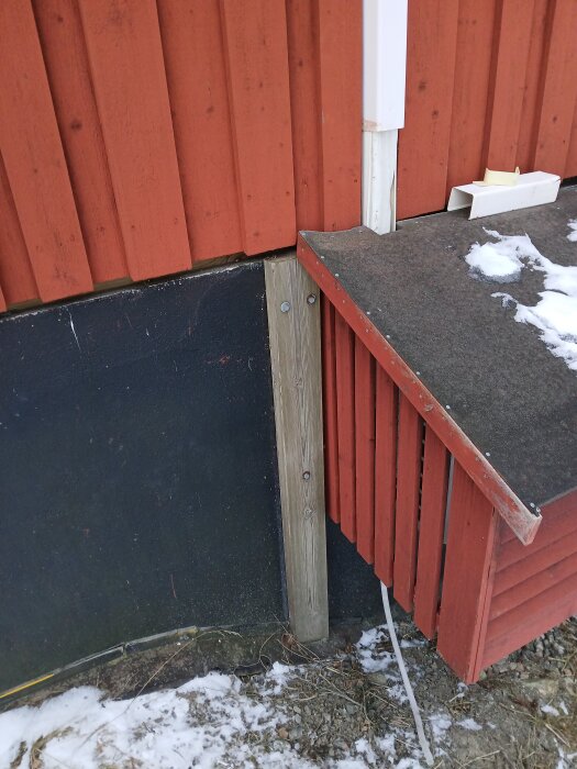 Rött trähus med luftvärmepumpsrör, vintertid. Dräneringsrör syns vid marken, täckt av delvis smält snö och is.