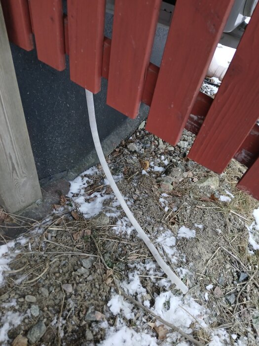 Dräneringsrör från luft-luft värmepump med is, delvis skyddat av röd träpanel, snö och grus på marken.