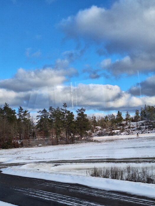 Snöig landskap med barrträd och klarblå himmel med moln. En asfalterad väg syns i förgrunden.