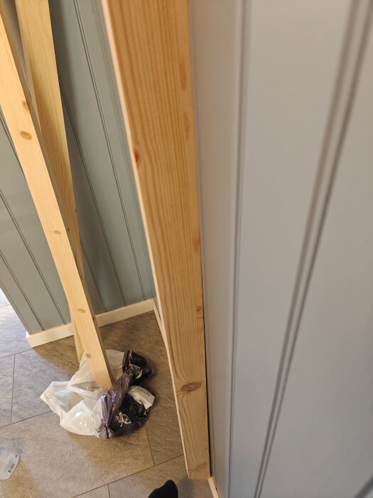 En lutande plankbit står lutad mot en ojämn vägg. Flera plankor och plastpåsar ligger på golvet bredvid.