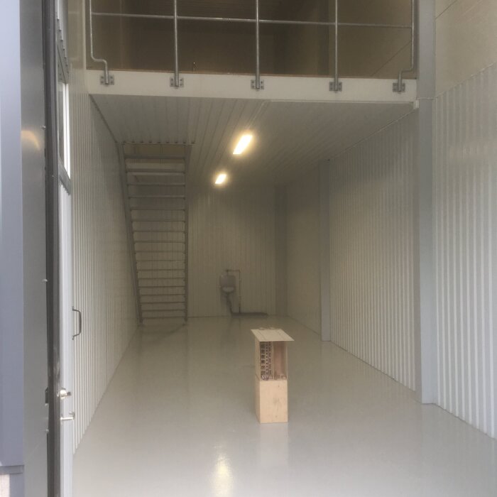 Tom lokal med vitmålat golv, trappa till loft och halvhöga väggar. Ett litet träbord står centralt i rummet framför en vägg.