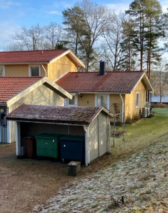 Gul enplansvilla med kaminens skorsten som når en meter ovanför taknocken, tvåvåningshus bredvid och grön och blå soptunna under carport.