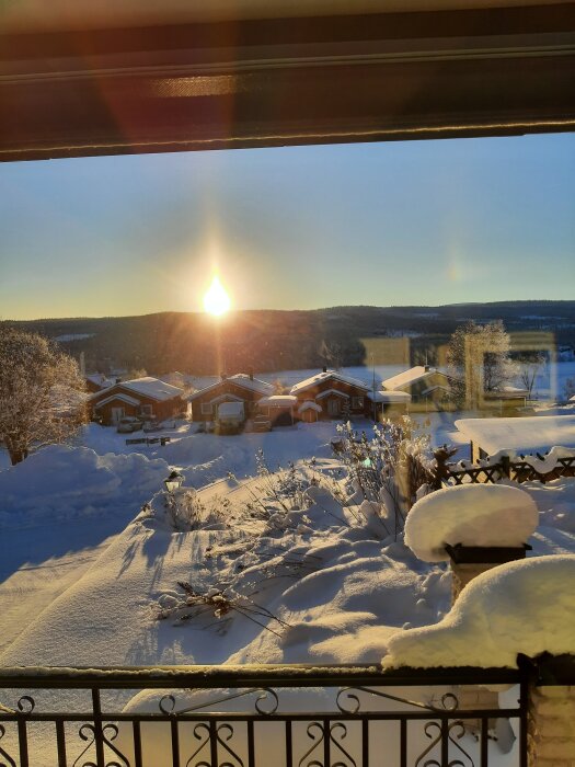 Snötäckt landskap med solen på väg ner bakom kullar, vy från en veranda med snöiga buskar och stugor i bakgrunden.