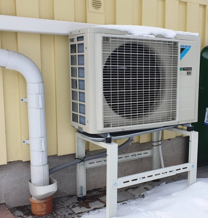 Utomhus luftvärmepump på väggen av ett gult hus med snö på. En uppvärmd dräneringsslang ansluten till pumpen.