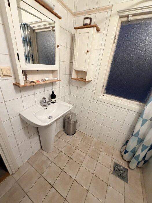 Badrum med handfat, spegelskåp, väggskåp, kaklade väggar och golv, avloppsbrunn och duschdraperi i bakgrunden.
