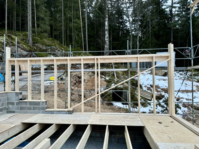 Byggarbetsplats med stomme av yttervägg i trä under konstruktion, omgiven av snötäckt mark och skog.