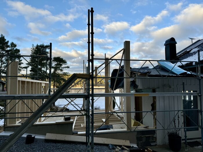 Bygge av ytterväggar och takstolar på en husrenovering bredvid en sjö, med träställningar och en delvis öppen struktur.