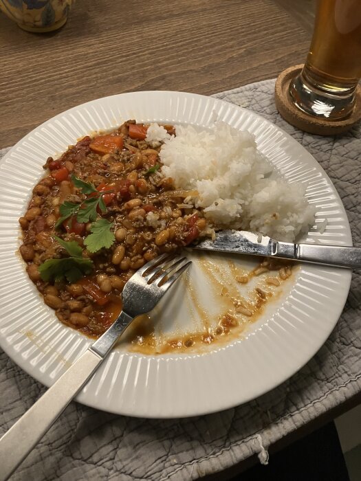 Tallrik med vita ris och köttgryta med bönor och paprika, garnerad med persilja, bredvid ett glas dryck på ett träbord.
