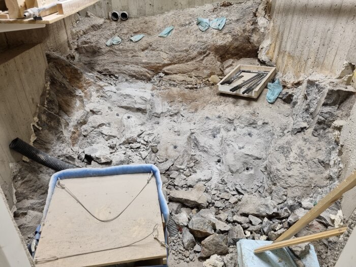 Utgrävd stenkällare med synliga borrhål i stenblock, omgiven av rubble och verktyg, inklusive trasiga borr i en träbehållare.
