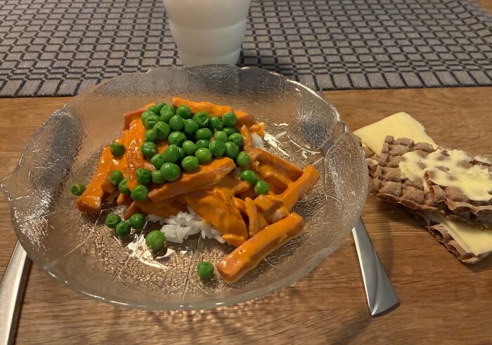 Tallrik med Korv Stroganoff och gröna ärtor på ris, bredvid knäckebröd med ost och dryck i bakgrunden.