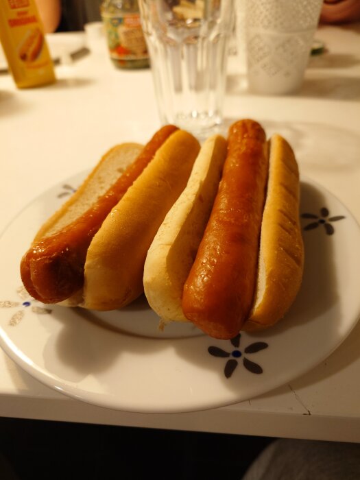 Två ugnsgrillade korvar i bröd på en tallrik, med senap och andra tillbehör synliga i bakgrunden.