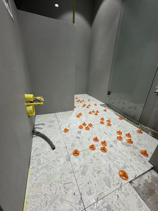 Kaklad golvyta med grå mönstrade plattor, orange kakelkilar är placerade på plattorna i ett rum under renovering.