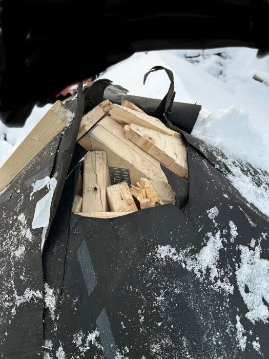 En hög med brutet virke täckt i snö på ett tak med svart takmaterial.