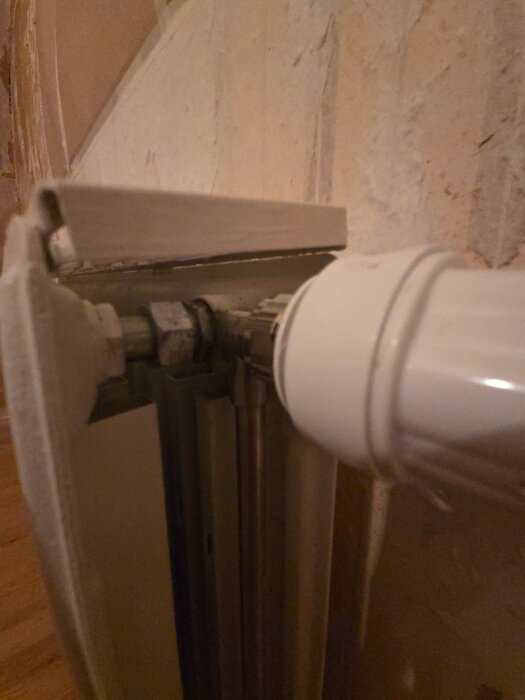 Närbild på en radiator med fokus på rörkopplingar och anslutningar, samt en vit ventil nära en vägg.