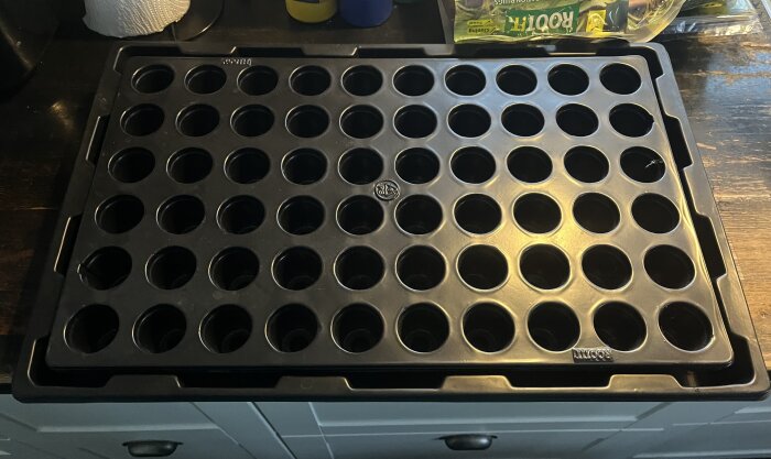 Svart odlingsbricka med flera små runda hål, placerad på en träbänk. Bakgrund med förpackning och hushållsartikel synlig.