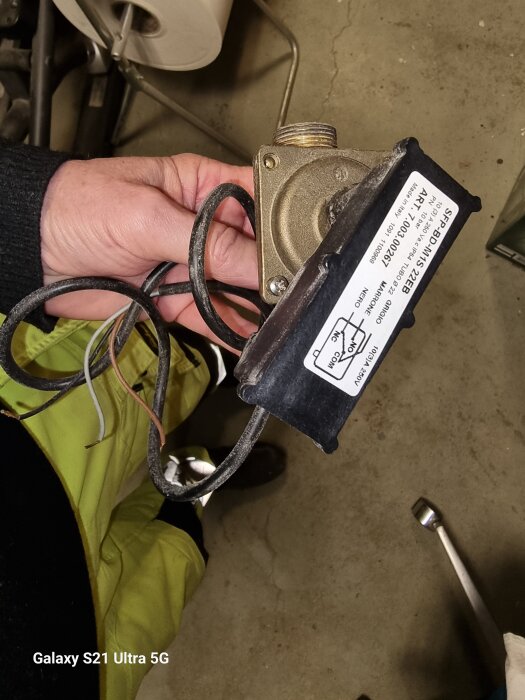 En hand håller en cirkulationspump eller ventil med elkablar och en produktetikett, placerad inomhus på ett grovt golv.