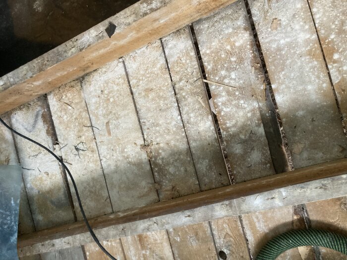Gammalt trägolv i sommarstuga under renovering, visar dammigt vitt pulver på plankorna mellan bjälkar.