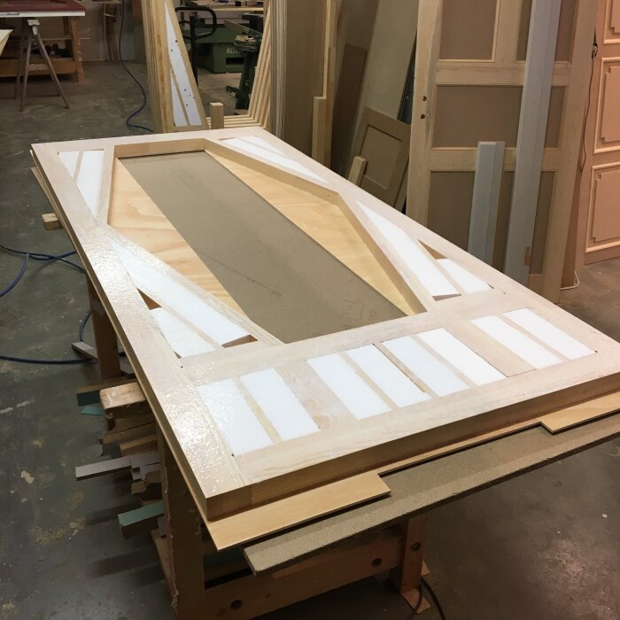 Träram med plywoodskiva i verkstad, under framställning av en dörr med sandwichkonstruktion. Material och verktyg synliga i bakgrunden.