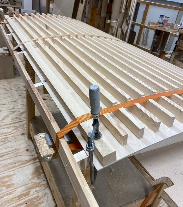 Träkonstruktion under limning, med stående ribbor fastspända på en plywoodskiva med spännband och tvingar i en verkstadsmiljö.