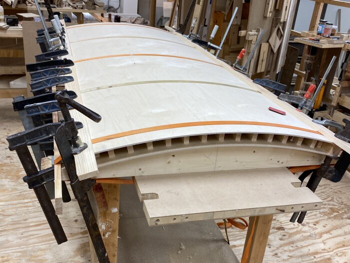 Formning av en svängd dörr med plywood och träribbor, fixerad med tvingar och spännband på en arbetsbänk i en snickeriverkstad.