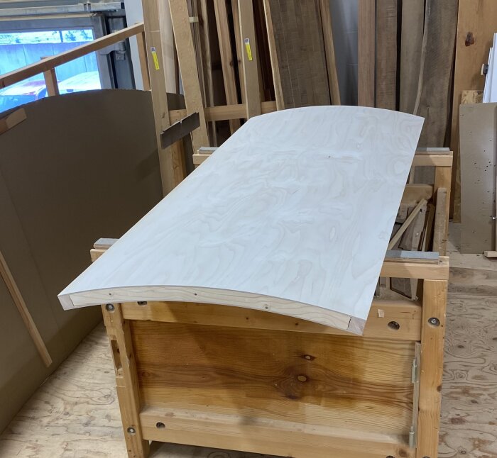 En svagt välvd träskiva av plywood placerad på en arbetsbänk i ett verkstadsmiljö, omgiven av andra trämaterial.