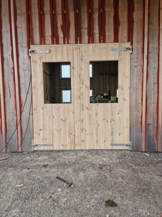 Träportar med två fönster under uppbyggnad, del av ett större byggprojekt med stående panel och synliga gångjärn. Ligghammare på marken framför.
