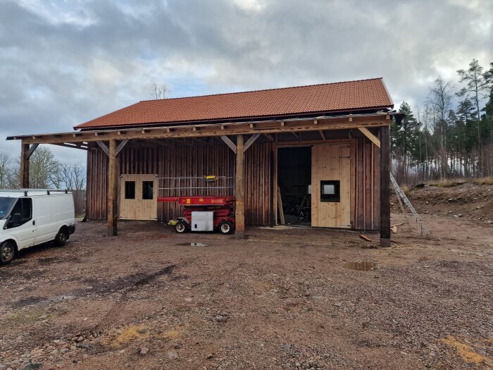 Träbyggnad med röda takpannor och dubbelportar, byggställning framför öppet garage, skog i bakgrunden.