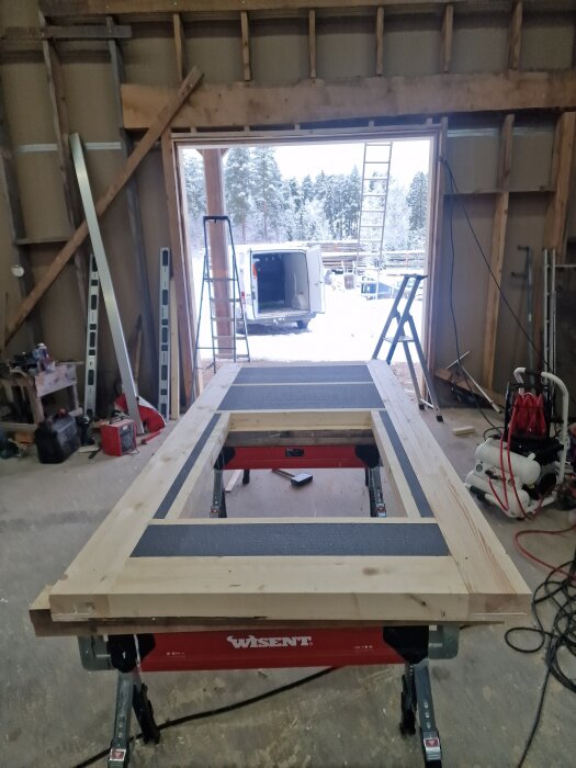 Träram till port under konstruktion i ett garage med verktyg runtom, en öppen dörr visar utsikten mot en gård med van och stegar.