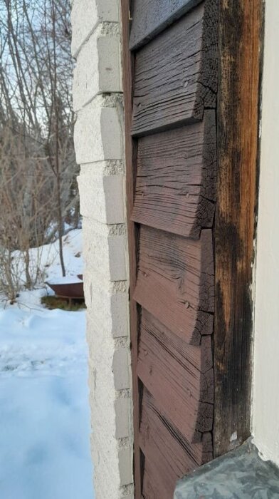 Närbild på träfasad med fjällpanel och synliga sprickor, liggande intill en murad stenvägg. Snö på marken i bakgrunden.