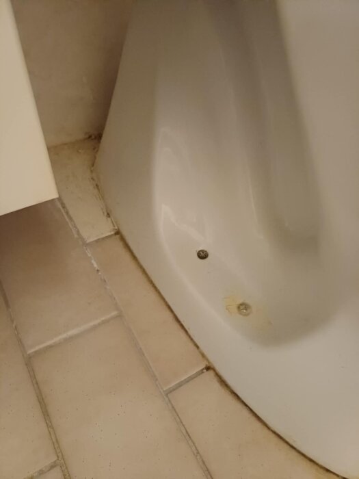 Toalettstolens botten vid beige kakelgolv, med synliga skruvhål och smutsfläckar.