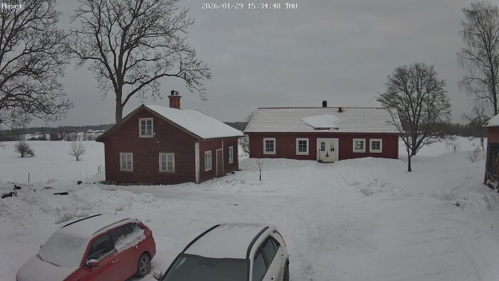 Snötäckt gårdsplan med två hus, ett rött och ett brunt, samt två parkerade bilar under vintern.